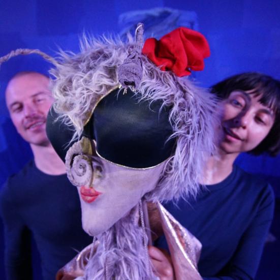 To skuespillere i en teaterkulisse står bag en dukke. 